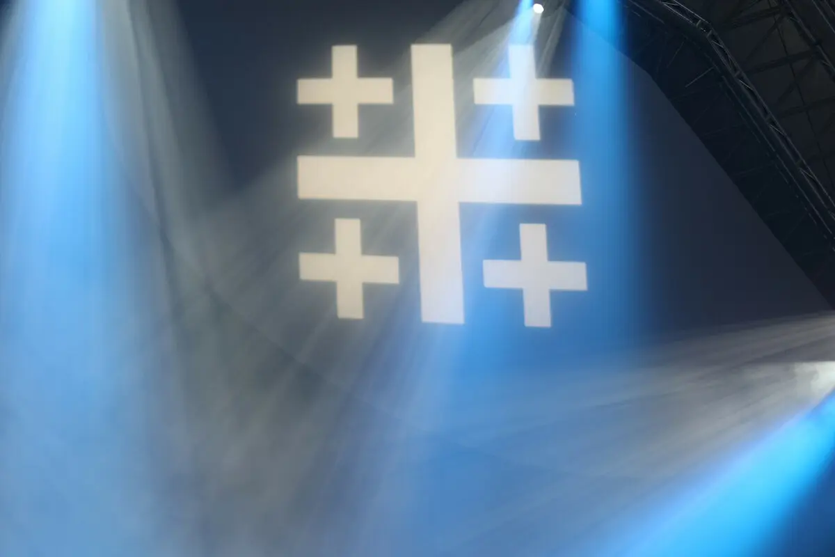 Ein Jerusalemkreuz wird in einer blauen Halle projiziert und Scheinwerfer bestrahlen es